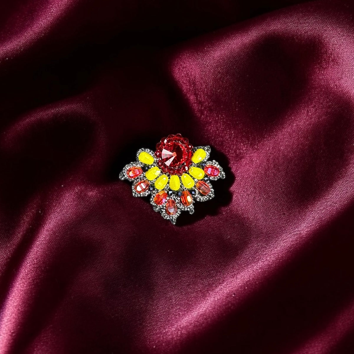 Spilla Fiammetta, brooch with red and yellow crystals on a burgundy fabric background.